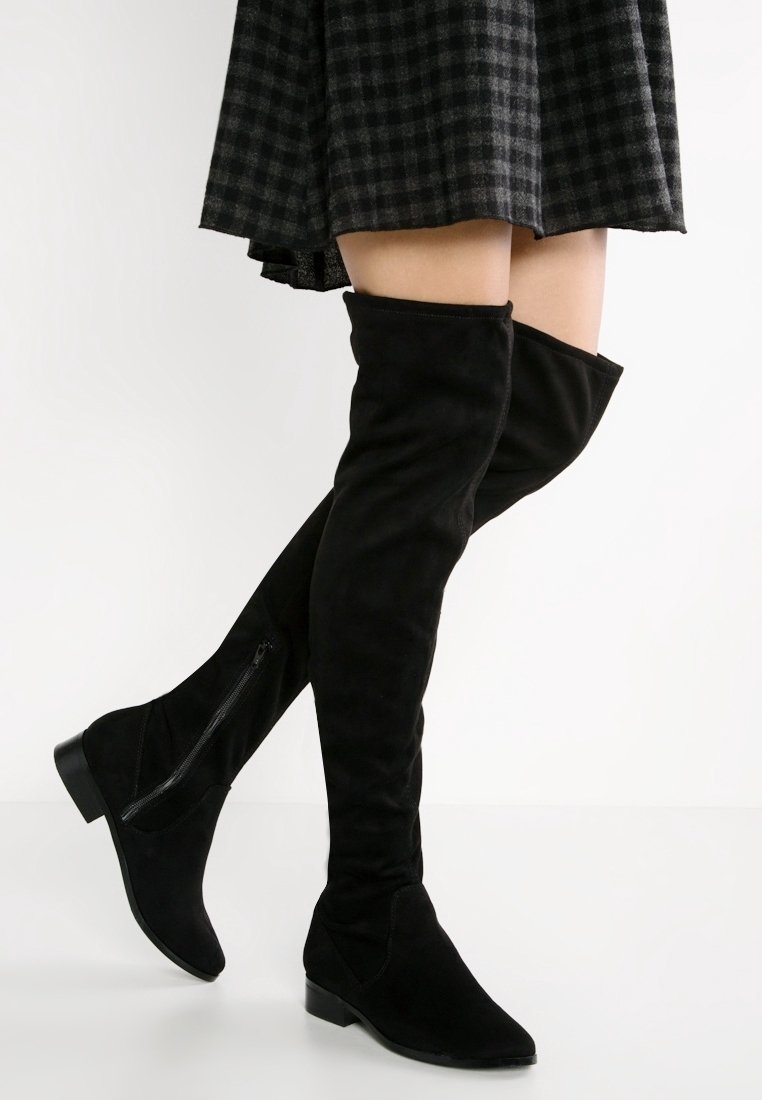 Černé nadkolenky vyrobené z měkkého semiše, s plochým podpatkem, bočním zipem a hladkou texturou. V doprovodu kostkované sukně.