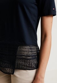 Námořnická modrá blůza s krátkými rukávy má krajkovou část na spodním okraji. Vidět je ruka, která spočívá na světlých kalhotách.