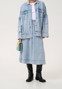 Henkilö, jolla on päällään vaaleansininen farkkutakki ja hame, valkoinen paita, mustat polvitason saappaat, ja joka pitää käsiss ään pientä ruskean ja sinisen väristä käsilaukkua.