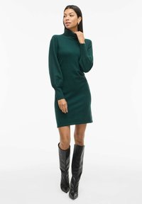 Vestido de punto verde con cuello alto, mangas abullonadas largas, forma ajustada y puños acanalados, combinado con botas altas de cuero negras.