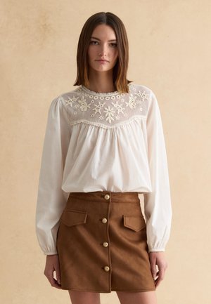 Mujer con una blusa blanca de manga larga con detalle de encaje floral en la parte superior y una falda marrón de ante con botones al frente, sobre un fondo beige liso.