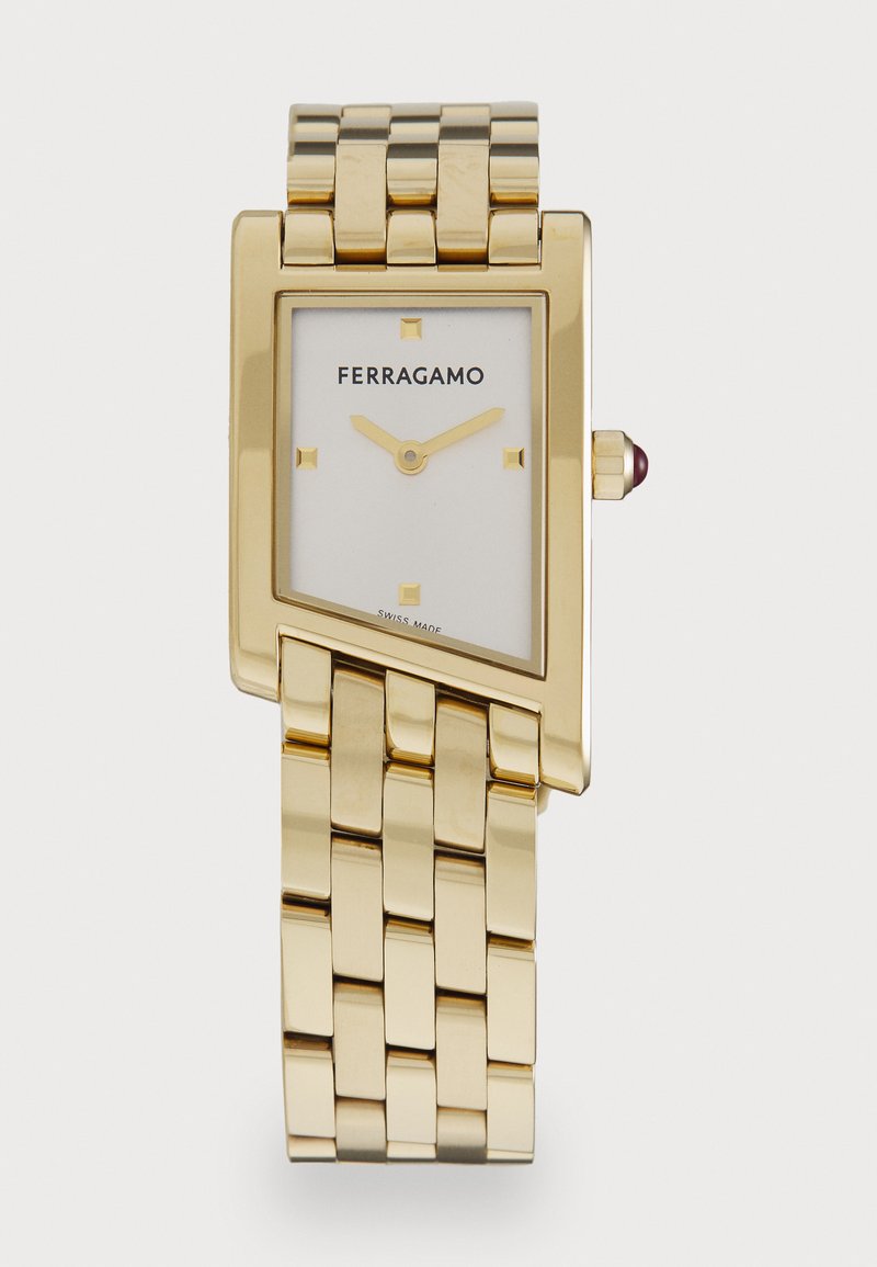 Kuldne ristkülikuline kell valge näo, kuldsed tunni näidikud ja tekstureeritud metallrand. Korona omab väikest punast aksenti.