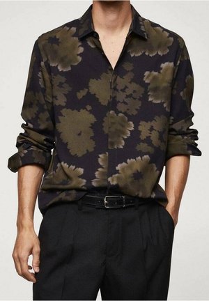 Mann trägt ein schwarzes Hemd mit großen olivgrünen Blumenmustern, hochgekrempelte Ärmel, in schwarze plissierte Hosen mit Gürtel gesteckt.