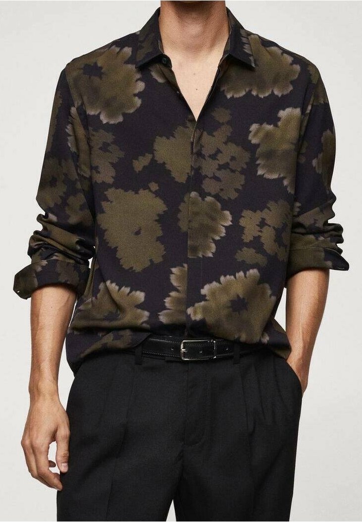 Homme portant une chemise noire à boutons avec de grands motifs floraux vert olive, manches retroussées, rentrée dans un pantalon noir plissé avec une ceinture.