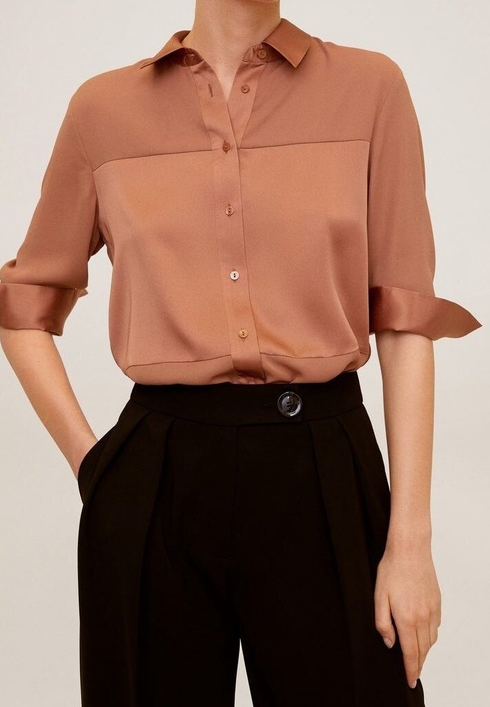 Femme portant une chemise à manches retroussées de couleur brun clair, boutonnée, rentrée dans un pantalon noir taille haute avec un grand bouton.