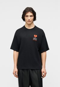 Νεαρός άνδρας με μεσαίου μήκους σκούρα μαλλιά που φοράει μαύρο υπερμεγέθες T-shirt με πορτοκαλί σπασμένη καρδιά και κείμενο στο αριστερό στήθος, μαύρο παντελόνι.