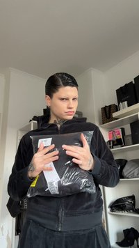 Svart emballasje som inneholder klær holdt foran en person som har på seg en svart fløyelshettejakke med glidelås. Gjennomsiktig plastfolie er synlig rundt varen.