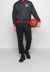 Sort bomberjakke med røde accenter og "BULLS STREET HOME" logo, parret med sorte sweatpants og holder en basketball.