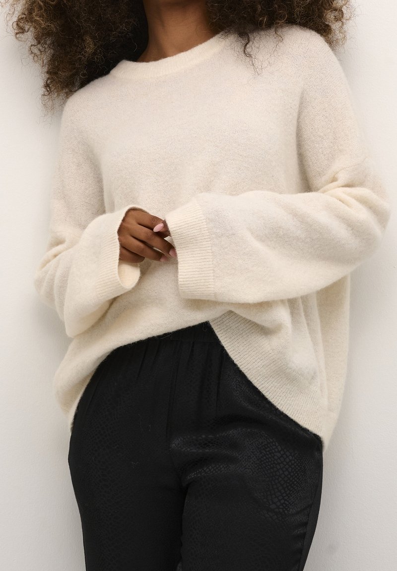 Personne portant un pull crème surdimensionné à manches longues et un pantalon noir texturé, les mains doucement jointes à la taille.