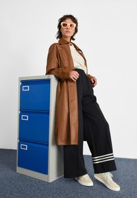 Casaco trench de pele castanha, blusa branca, calças largas pretas com riscas e sapatilhas bege. Modelo posa contra um armário de arquivos azul.