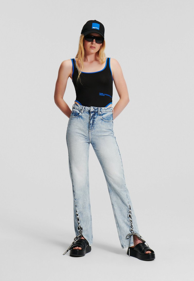 Zwarte body met blauwe afwerking, lichte jeans met uitsnijdingen aan de onderkant, zwarte platform sandalen en een zwarte pet met blauw logo.