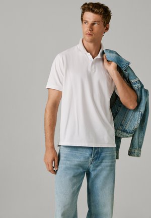 Weißes Poloshirt mit Kragen und kurzen Ärmeln, kombiniert mit hellblauen Jeans. Eine ausgewaschene blaue Jeansjacke wird über der Schulter getragen.