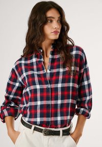 Κόκκινο, ναυτικό και λευκό καρό πουκάμισο με γιακά που κουμπώνει, μακριά μανίκια και χαλαρή εφαρμογή, φορεμένο μερικώς τυλιγμένο μέσα σε ανοιχτόχρωμο παντελόνι.