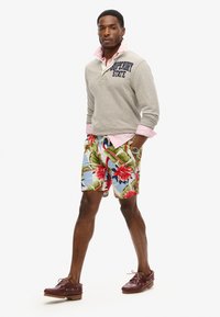 Szary polarowy sweter z napisem "Superdry State", w połączeniu z kolorowymi kwiecistymi szortami kąpielowymi. Ciemnobrązowe buty żeglarskie dopełniają całość.
