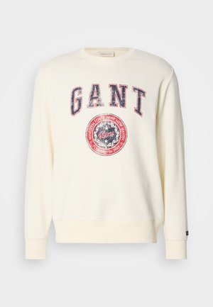 Sweatshirt creme com mangas longas, gola redonda e um gráfico do logo GANT desgastado em vermelho e azul. Textura de tecido suave.