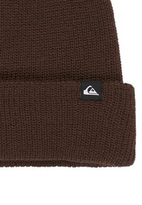 Brown knit beanie with a folded cuff. Features a black label with a white logo on the side. Texture is ribbed and slightly loose.