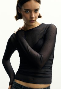 Fekete, hosszú ujjú felső áttetsző anyagból, apró díszes pöttyös részletekkel és testhezálló szabással. Simább textúra, gyűrt díszítéssel.