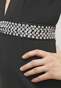 Černé šaty s stříbrnou květinovou výšivkou na pase. Hladký materiál, přiléhavá silueta a hluboký výstřih.