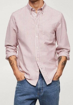 Homme portant une chemise à rayures roses et blanches à boutons, manches retroussées, jeans bleus, mains dans les poches, tatouages visibles sur les bras.