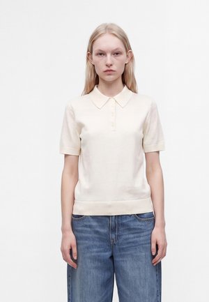 Jeune femme debout portant un polo crème à manches courtes et un jean bleu devant un fond blanc uni.