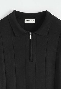 Polo noir en tissu texturé, avec une fermeture éclair à l'avant, un col côtelé et des détails de couture verticale. Étiquette de la marque visible à l'intérieur.