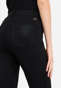 Pantalon noir avec une poche arrière texturée et perforée au design hexagonal, agrémentée de quincaillerie dorée à la taille pour plus de détails.