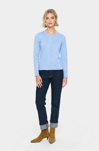 Cardigan côtelé bleu clair avec col rond et fermeture à boutons. Associé à un jean bleu foncé et des bottines beiges, mettant en avant un look décontracté.