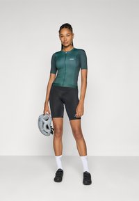 Tricou de ciclism cu mânecă scurtă, de culoare verde închis, cu fermoar pe față, asortat cu pantaloni scurți cu captuseală negri și o cască de ciclism gri.