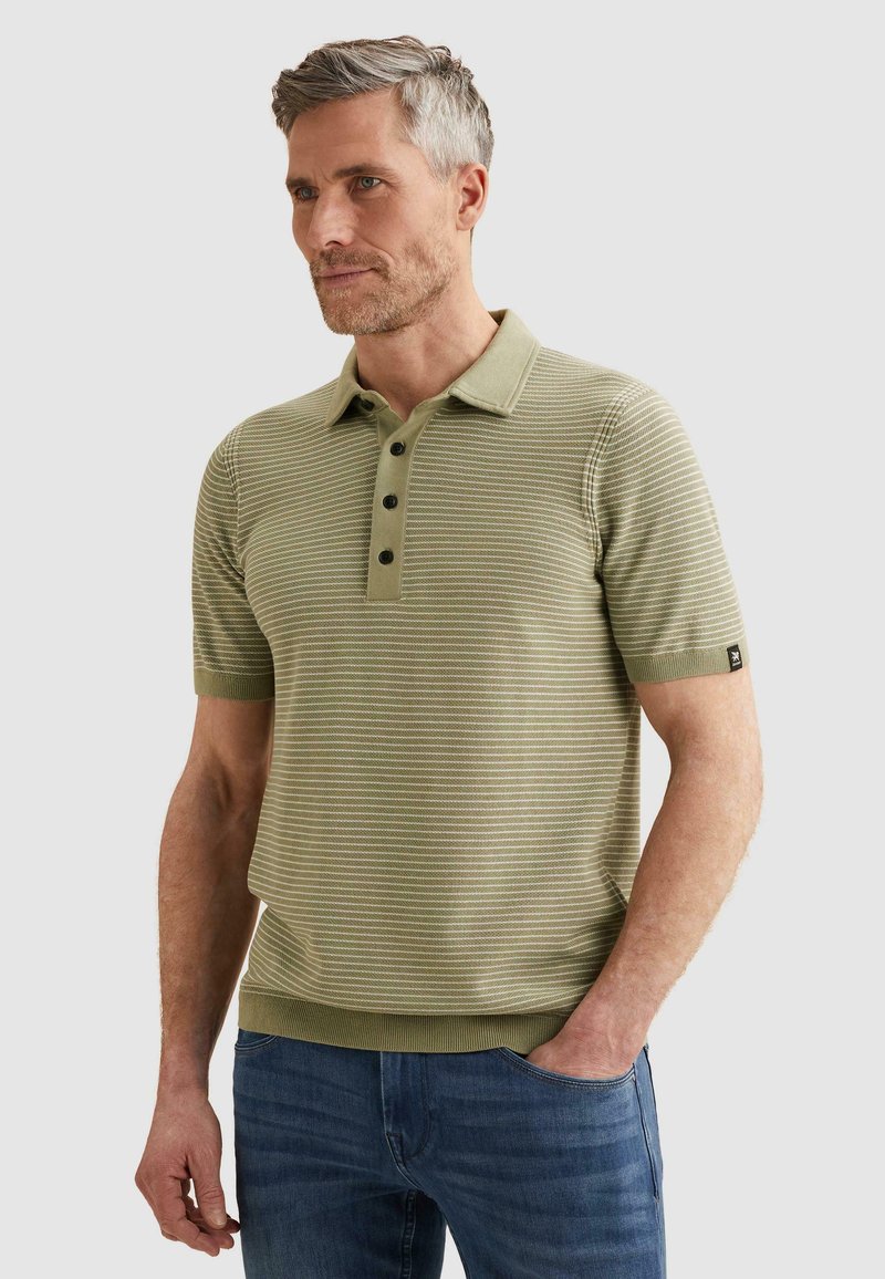Homme aux cheveux gris courts, portant un polo rayé vert olive clair et un jean bleu, debout avec une main dans la poche.