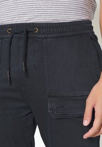 Musta, joustavavyötäröiset juoksuhousut, joissa on kiristysnauha, sivutaskut ja saumatut korostukset sisältävä cargo-tasku. Pehmeä kangaspinta.