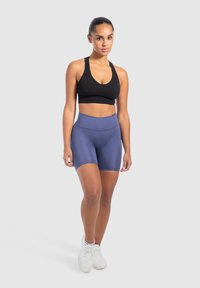 Zwarte sportbeha en blauwe high-waisted shorts, beide strak passend en gemaakt van rekbare stof. Model draagt witte sportschoenen, minimale achtergrond.