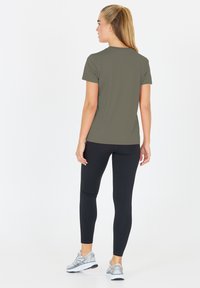 Olivgrön kortärmad träningsskjorta i kombination med svarta leggings och silverfärgade lö sneakers, sedd bakifrån mot en vit bakgrund.