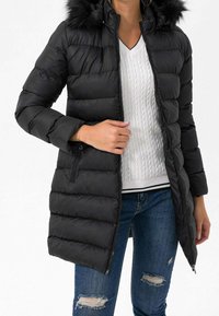 Sort pufferjakke med en pelsforet hætte, lynlås foran og ribbede detaljerne i kanten. Sammen med en hvid kabelstrikket sweater og slidte blå jeans.