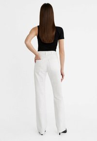 Pantaloni bianchi a vita alta e svasati con una texture liscia, due tasche posteriori e un top aderente nero. Modella vista da dietro.