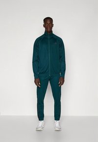 Mjukisdräkt i mörk teal, gjord av slätt, stretchigt material. Innehåller en jacka med dragkedja och avsmalnande byxor med svarta sidostripes. Accentuerad med en logotyp.