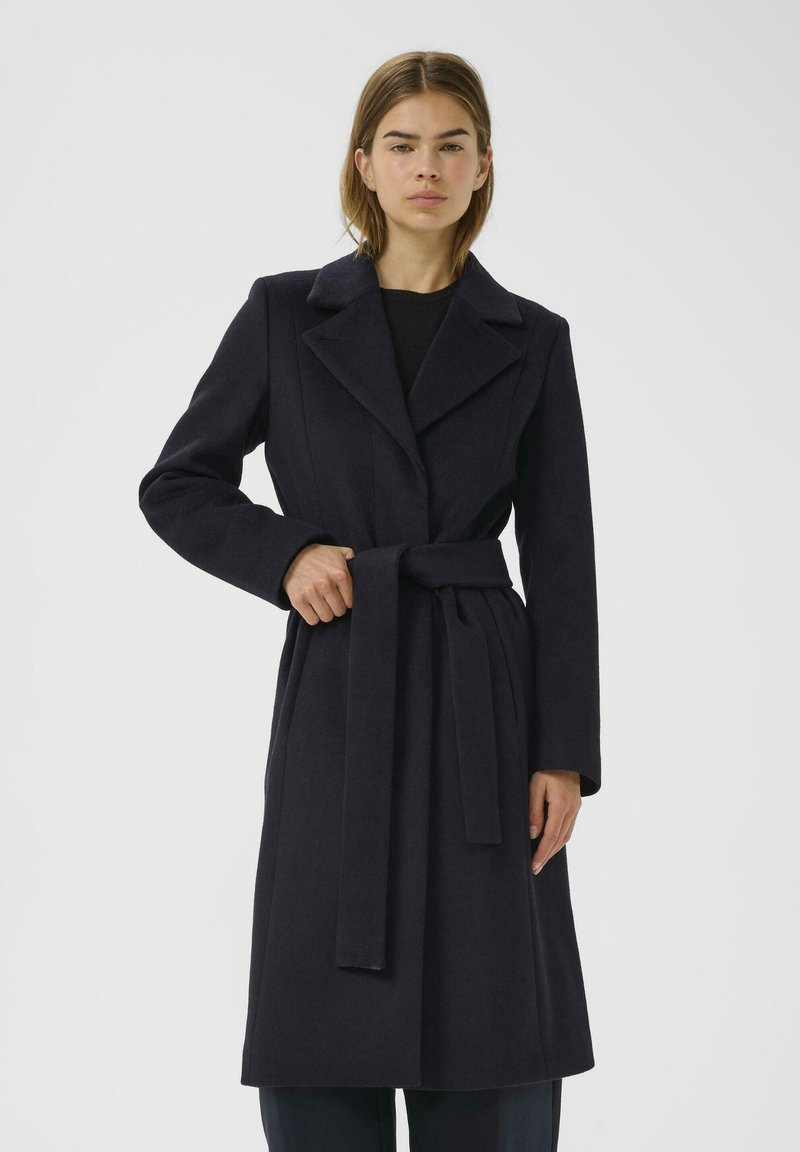 Cappotto trench in lana blu navy con una vestibilità su misura, colletto a punta e vita con cintura. Maniche lunghe e texture liscia con dettagli minimi.
