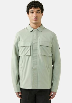 Joven con cabello rizado que lleva una chaqueta-camisa verde clara con dos grandes bolsillos en el pecho, de pie frente a un fondo blanco liso.