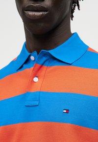 Κοντινό πλάνο ενός ατόμου που φοράει ριγέ μπλε και πορτοκαλί polo με ένα μικρό ορθογώνιο κόκκινο, λευκό και ναυτικό λογότυπο στο στήθος.