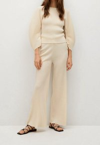 Top en tricot beige à côtes avec des manches bouffantes, associé à un pantalon large assorti. Le mannequin porte des sandales noires à lanières. Tissu léger et texturé.