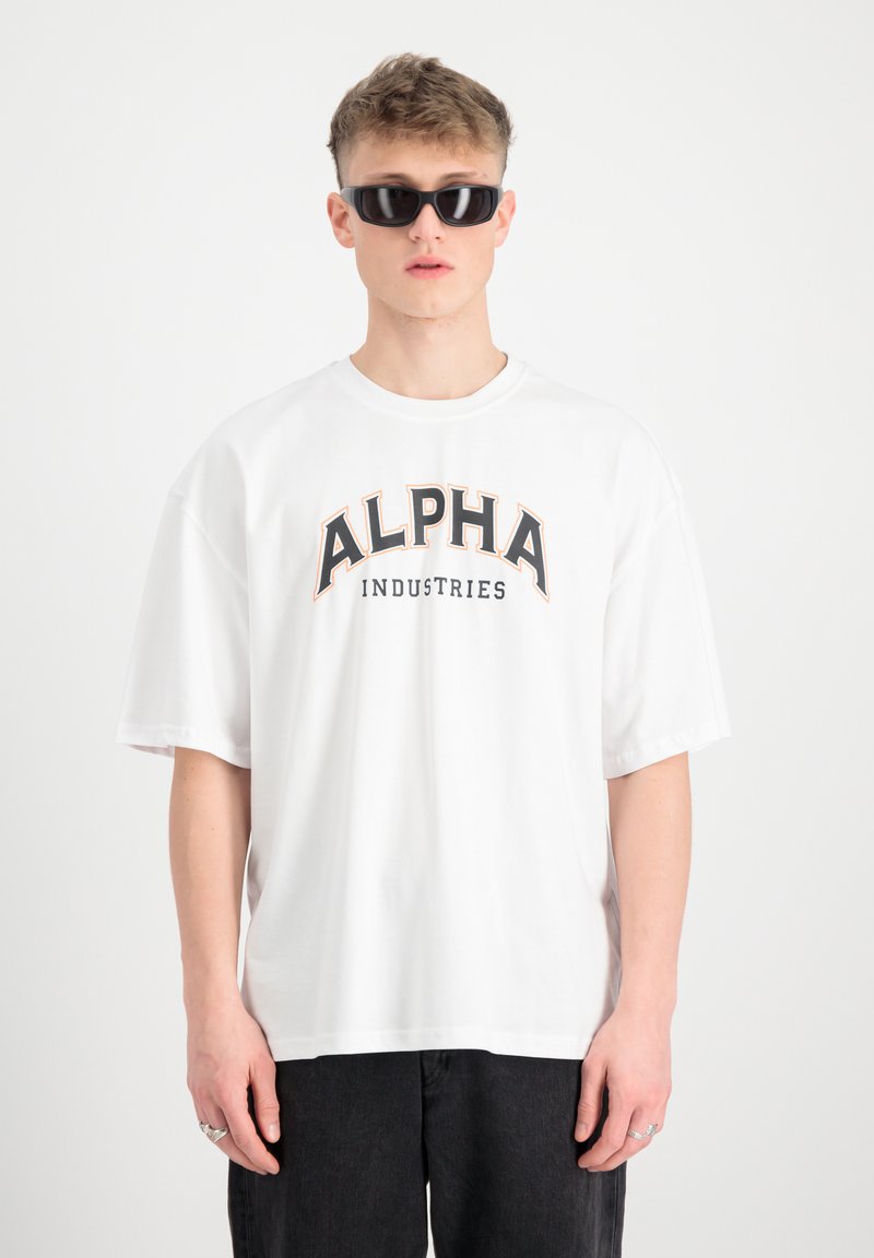 Bela oversize bomba majica iz bombaža z grafiko "ALPHA INDUSTRIES" v črni in oranžni barvi. Model nosi črne sončne očala. Preprost dizajn, kratki rokavi.
