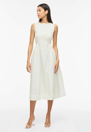 Ženska, ki nosi belo midi obleko brez rokavov z navpičnimi detajli šivov, stoji in gleda levo na enobarvni beli podlagi.