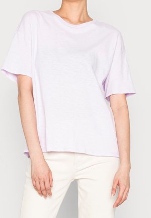 Person iført en lyse-lilla kortærmet løs T-shirt og offwhite bukser, stående med armene afslappet langs siderne.