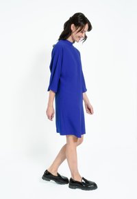 Kraljevo plava haljina širokog kroja s visokim dekolteom i rukavima do tri četvrtine, uparena s crnim cipelama na punu petu koje imaju debeli potplat.