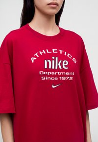 Νέος που φοράει άνετο κόκκινο μπλουζάκι Nike με λευκό κείμενο "Athletics Nike Department Since 1972" και μικρό λογότυπο Nike.