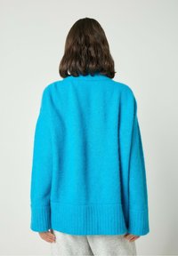 Garais piedurknēs džemperis dzīvā zilā krāsā, ar teksturētu dzijas rakstu un ribotu apakšmalu. Siluets ir pārāk liels un ar augstu apkakli.