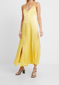 Robe maxi en satin jaune avec un décolleté en V, fermeture à boutons sur le devant et fentes latérales. Présente des fines bretelles et une silhouette fluide.