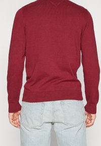 Maglione in maglia bordeaux con scollo a V, maniche lunghe e polsini e orlo a costine. Indossato con jeans azzurri chiari, che mostrano un outfit casual.