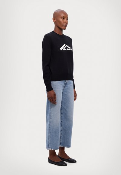 Zwarte trui met het "ICON" logo in het wit, gecombineerd met lichtblauwe wijde jeans. Model draagt zwarte platte schoenen; alle kledingstukken hebben een zachte textuur.