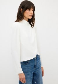 Blusa bianca a maniche lunghe con colletto alto e dettagli plissettati. Tessuto liscio, vestibilità leggermente più ampia, abbinata a jeans blu con decorazioni.