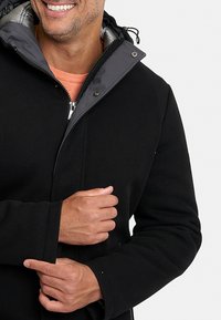Chaqueta negra con capucha y cremallera frontal, con un forro interior gris y textura acanalada en las mangas. Cordon ajustable en la capucha.
